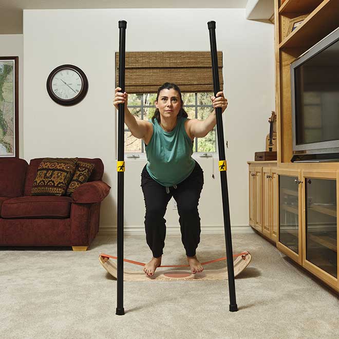 Woman using two balance poles while squating on a sunbeam slackboard in a living room.
